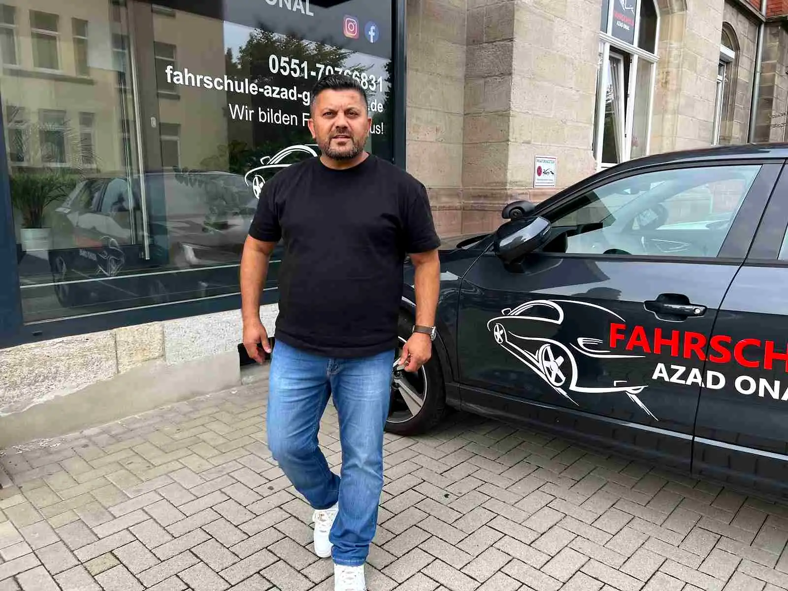 Azad Onal vor dem Ladenlokal der Fahrschule AZAD mit beschriftetem Fahrschulwagen